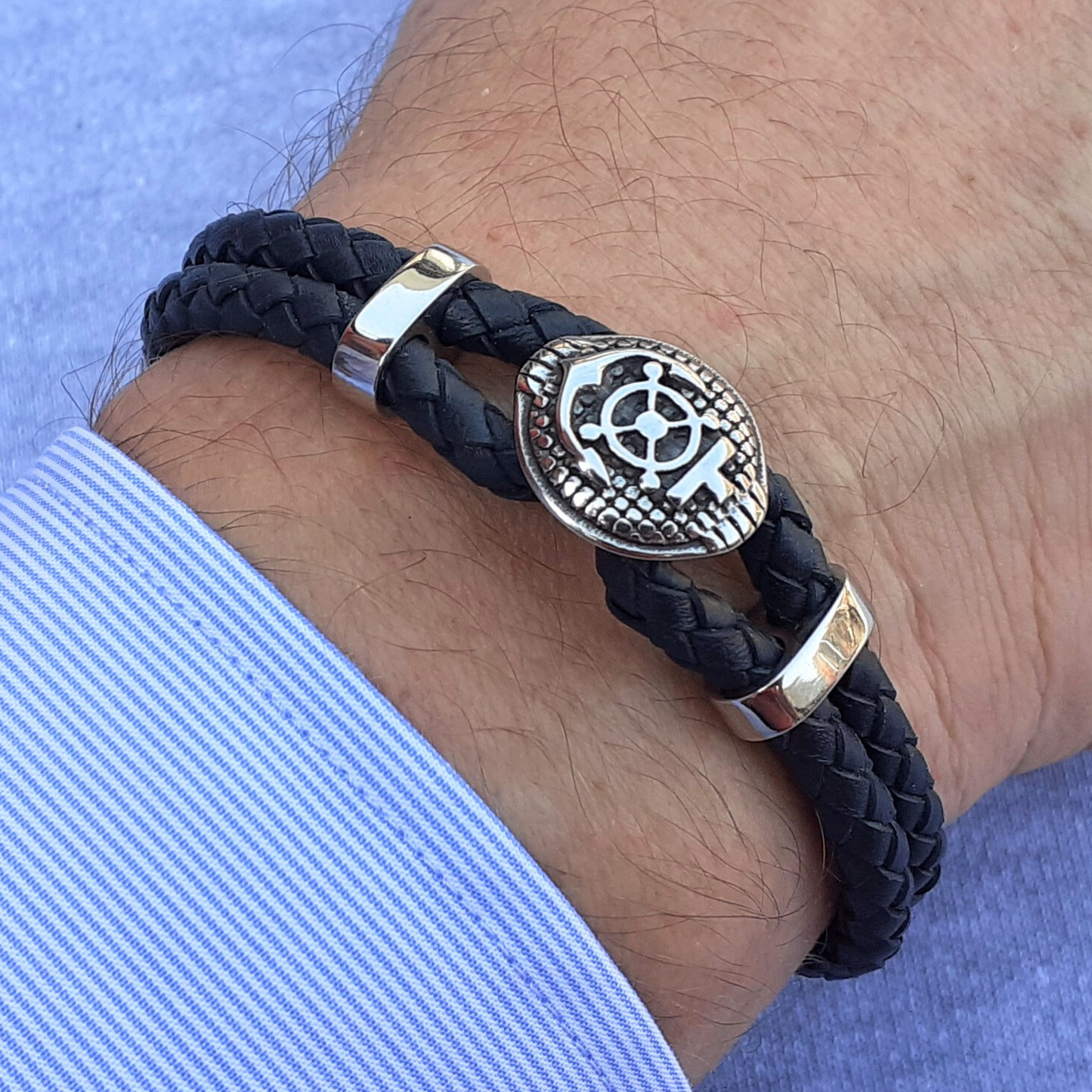 Black braided bracelet with a stainless steel compass and anchor emblem on a wrist against a light blue background.