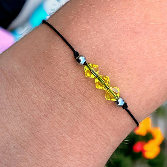 A bracelet with yellow citrine crystal beads and stainless steel beads, tied with a black macrame knot, worn on a wrist with a colorful background.