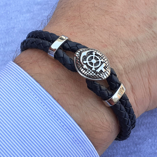 Black braided bracelet with a stainless steel compass and anchor emblem on a wrist against a light blue background.