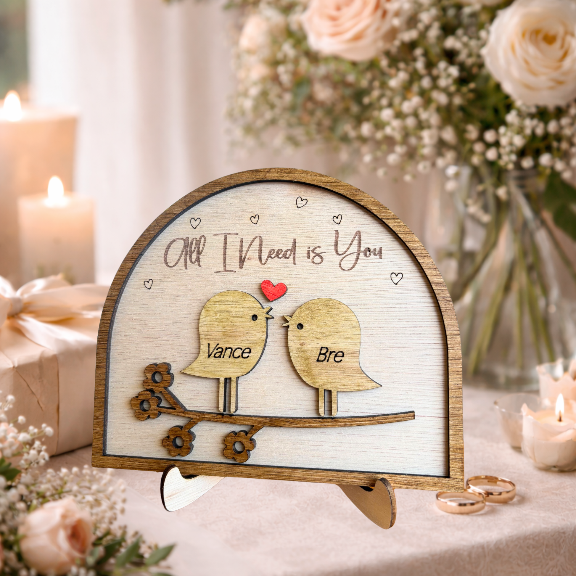 Wooden plaque with two love birds and 'All I Need is You' text, surrounded by flowers and candles.