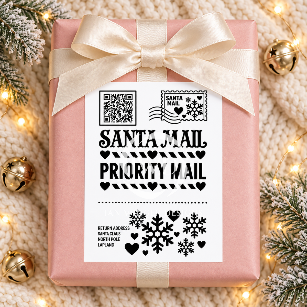 A pink gift-wrapped box tied with a cream satin ribbon, featuring a white Santa Mail Priority Mail label with black text and holiday graphics, styled on a knitted cream surface with warm string lights and gold bells.